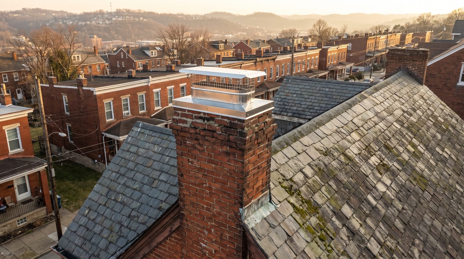 Brick residential chimney with a stainless steel cap on a Pittsburgh rooftop at dusk, city hillside behind
